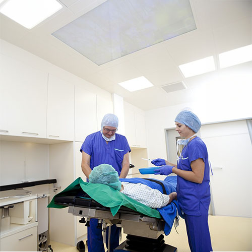 Ein Behandlungsraum mit zwei Ärzte im blauen Kiteln, Mundschutz und OP-Haube sowie ein Patient in weißen Kitel und OP-Haube. Eine Ärztin hält eine blaue Nierenschale mit Material .jpg